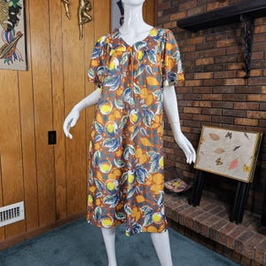 May include: A knee-length dress featuring a vibrant fruit print with oranges, lemons, and green leaves set against a brown background. The garment has short, loose sleeves, a rounded neckline, and a relaxed, flowing silhouette that falls straight.