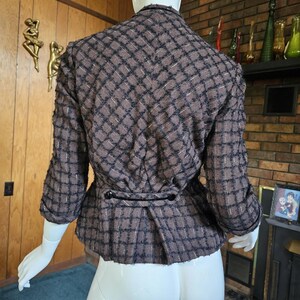 May include: Brown and black plaid blazer with three-quarter sleeves. The jacket features a fitted waist with two black buttons and a textured fabric. The back of the jacket is visible, showcasing the tailored design and pattern.