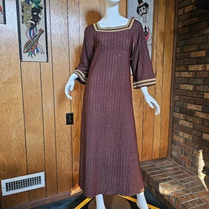 May include: A long, brown dress with gold trim around the neckline and sleeves. The dress has a vertical striped pattern and a loose, flowing silhouette.