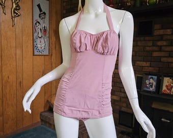 May include: A vintage-style, light pink halter-neck swimsuit displayed on a mannequin. The one-piece suit features a ruched bodice and sides, with a matching bottom. The background includes a wooden wall and vintage decor.