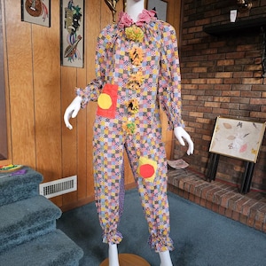 May include: A colorful clown jumpsuit with a patchwork pattern in various colors. The costume features a ruffled collar, pom-poms, and large, round patches. The long-sleeved jumpsuit has ruffled cuffs and is displayed on a mannequin.