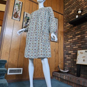 May include: A long-sleeved dress on a mannequin. The dress has a cream background with a repeating pattern of blue, brown, and maroon floral shapes. The dress is knee-length and has a loose, comfortable fit. The mannequin is standing in front of a wood-paneled wall.