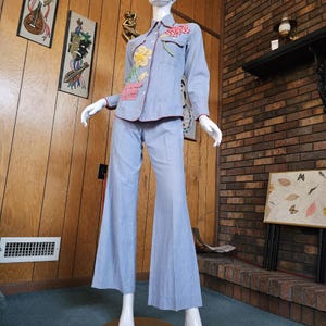 May include: A light blue denim two-piece outfit on a mannequin. The long-sleeved shirt has floral and gingham patches with red trim. The matching pants are flared. The outfit is vintage.