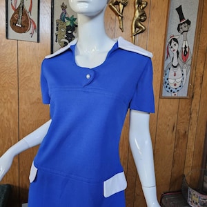May include: A vintage blue dress with white collar and pocket accents. The dress features short sleeves and a single button closure at the neckline. The dress is displayed on a mannequin.