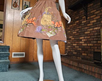 May include: Brown pleated skirt with a printed design featuring a basket, yarn, and knitting needles. The skirt is displayed on a mannequin. The design includes orange, purple, and green accents. The skirt has a wide waistband and a flared shape.