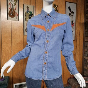 May include: A light blue denim shirt with long sleeves and a pointed collar. The shirt features a brown leather-like accent across the chest and wooden buttons down the front. The shirt is displayed on a mannequin.