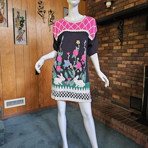 May include: A dark gray knit dress with a pink and white diamond pattern on the shoulders. The dress features a floral and bunny design with pink, blue, and yellow flowers. The hemline has a black and white checkered pattern.