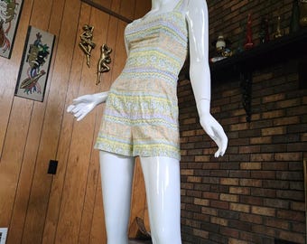 May include: A vintage romper in pastel colors of yellow, lavender, and peach, displayed on a mannequin. The romper features a square neckline, thin straps, and a patterned design. The shorts are short and the fabric appears to be a woven material.