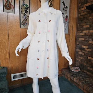 May include: Cream-colored long cardigan sweater with a collared neckline, button closure, and two front pockets. The sweater features embroidered floral and insect designs in red, pink, and purple. The sleeves and front panels have a diamond pattern.
