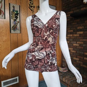 May include: A vintage one-piece swimsuit with a V-neck and a floral pattern in shades of brown, beige, and gray. The swimsuit is sleeveless and has a ruched waist detail. The garment is displayed on a mannequin.