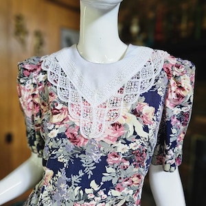 May include: A vintage floral dress with a white lace collar. The dress features a navy blue background with a pattern of pink, purple, and cream-colored flowers. The short sleeves are puffed.
