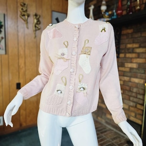 May include: A pink knitted cardigan with a button-up front and embroidered Christmas stocking designs. The stockings are decorated with white and gold accents.