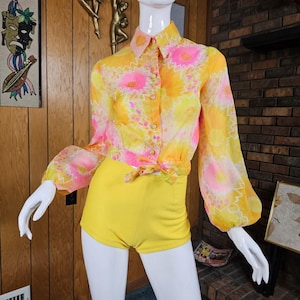 May include: A long-sleeved button-down shirt featuring a vibrant yellow and pink floral pattern with a tie-front waist, paired with solid yellow high-waisted shorts. The shirt has a pointed collar and full sleeves with gathered cuffs on a mannequin.