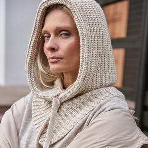 May include: A beige knit hood and scarf combination with a drawstring closure. The hood is worn over a light gray sweater and a beige puffer jacket. The outfit is neutral-toned and suitable for cold weather.