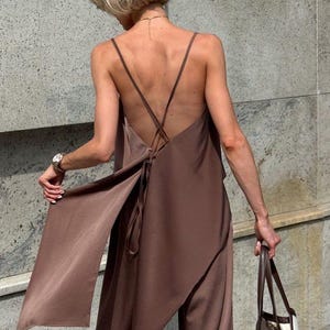 May include: A brown satin jumpsuit with a draped top and wide-leg pants. The top features thin straps and a low back with a tie detail. A white tote bag with brown handles is held in the right hand. The outfit is worn outdoors.