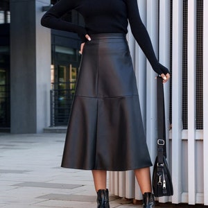 Pu&ograve; includere: Una donna indossa un maglione nero a collo alto, una gonna di pelle nera, un berretto da baseball nero con le lettere "NY" e stivaletti neri. Porta una borsa a tracolla nera.