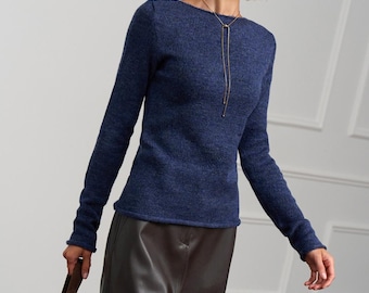 May include: A long-sleeved, dark blue knit sweater with a boat neckline, paired with a dark brown leather skirt and a matching brown suede handbag. The outfit is accessorized with a thin gold-toned lariat necklace and gold hoop earrings.