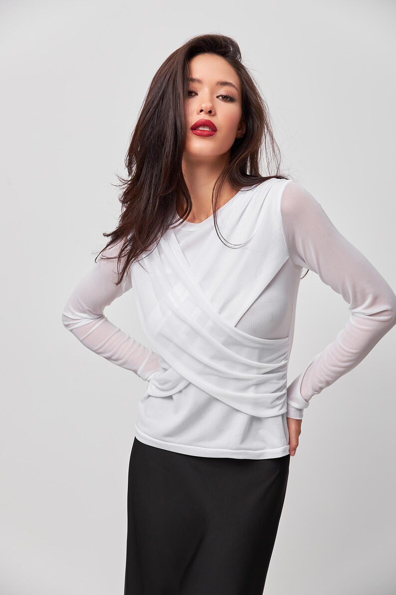 May include: A white long-sleeved shirt with a criss-cross design in the front. The shirt is made of a sheer fabric and has a relaxed fit.