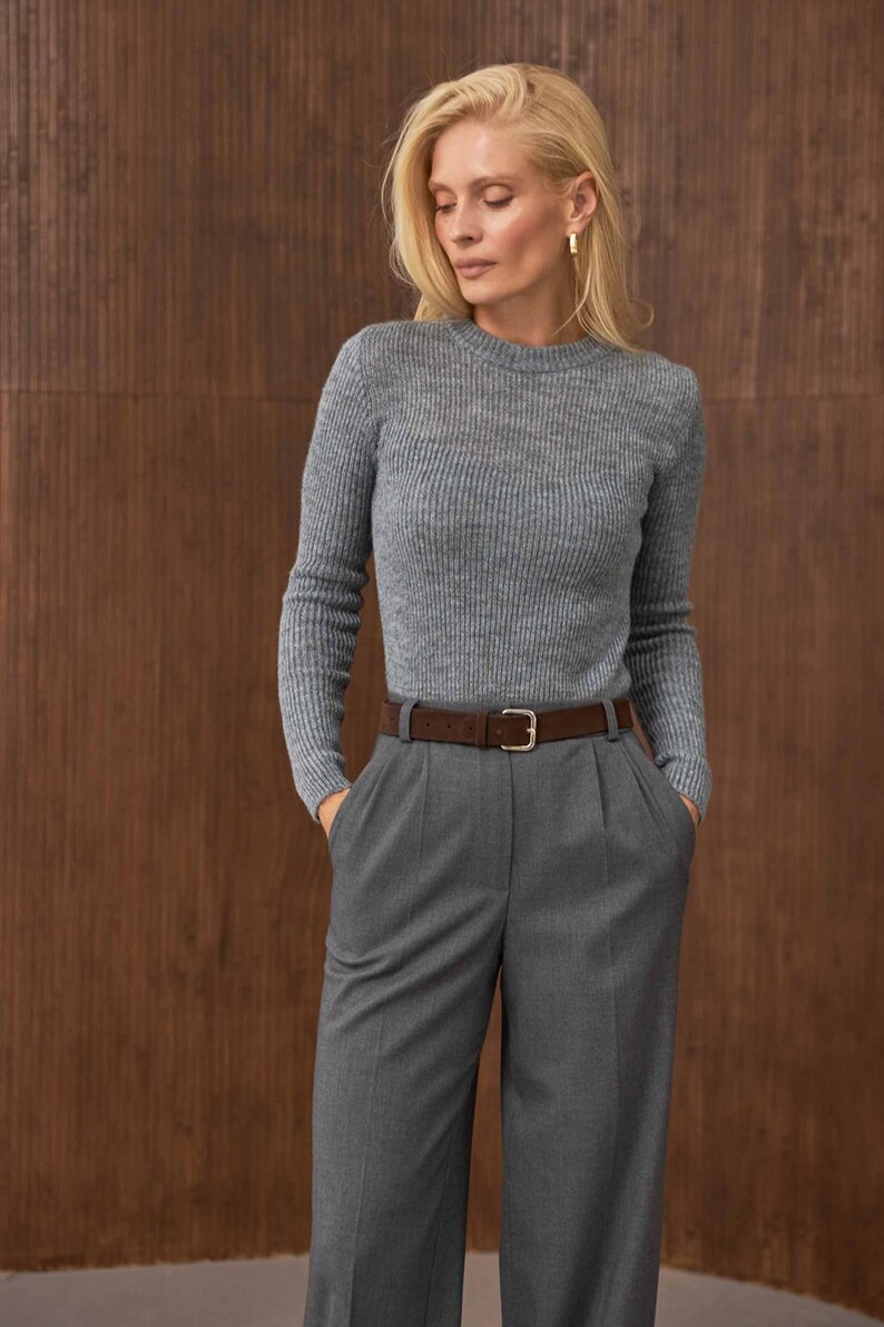 May include: A woman wearing a gray ribbed sweater, gray trousers, and a brown belt with a silver buckle. The outfit is a monochromatic ensemble, with the sweater tucked into the high-waisted pants. The background is a brown wooden wall.