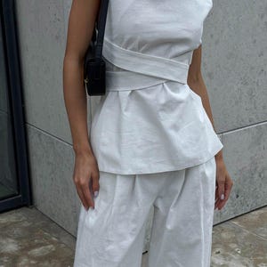 May include: A white linen top with a wrap-style design and a matching pair of wide-leg pants. The top has a scoop neck and sleeveless design. The pants have a high waist and a relaxed fit.