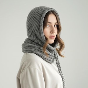 May include: A gray knitted hood with a textured pattern. The hood is designed to cover the head and neck, featuring a cowl neck and braided ties. The item is made of a soft, warm material, perfect for cold weather.