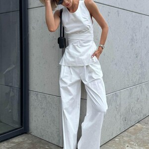 May include: A white sleeveless top with a wrap detail and a matching pair of wide-leg pants. The outfit is styled with a small black handbag and sunglasses.
