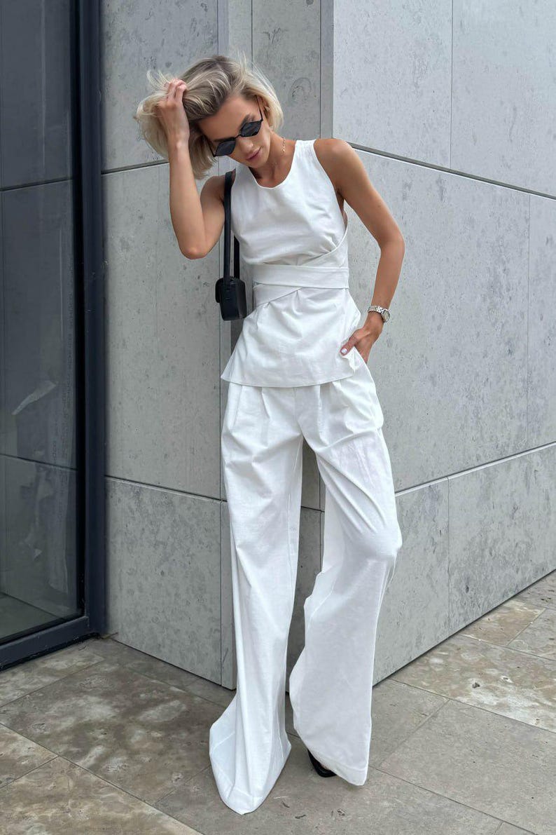 May include: A white sleeveless top with a wrap detail and a matching pair of wide-leg pants. The outfit is styled with a small black handbag and sunglasses.