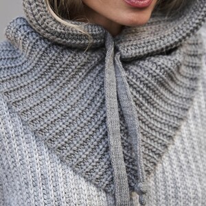 May include: Close-up of a gray knitted hood and cowl. The hood is attached to a sweater, with a drawstring closure. The knitwear has a ribbed texture and is in shades of gray.