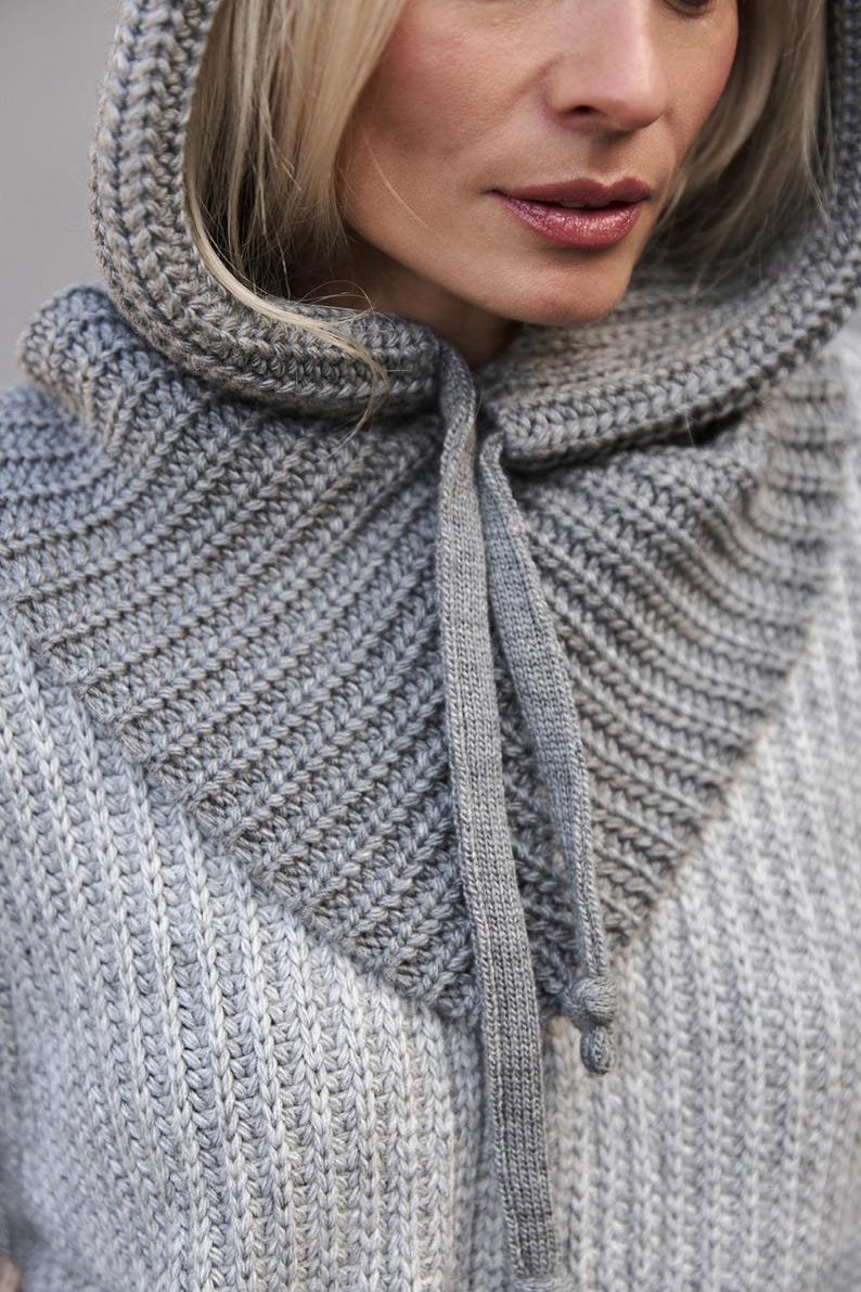 May include: Close-up of a gray knitted hood and cowl. The hood is attached to a sweater, with a drawstring closure. The knitwear has a ribbed texture and is in shades of gray.