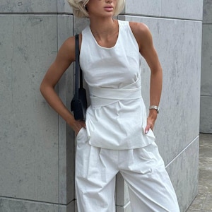May include: A woman wearing a white sleeveless top with a knotted detail at the waist and matching white wide-leg pants. She is wearing black sunglasses and a small black purse.