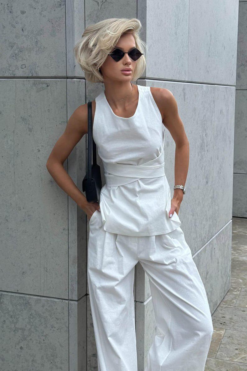 May include: A woman wearing a white sleeveless top with a knotted detail at the waist and matching white wide-leg pants. She is wearing black sunglasses and a small black purse.