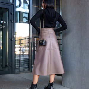 Pu&ograve; includere: Una donna indossa un maglione nero con cappuccio e una gonna di pelle marrone. Porta una piccola borsa nera con una tracolla a catena dorata. Indossa stivaletti neri.