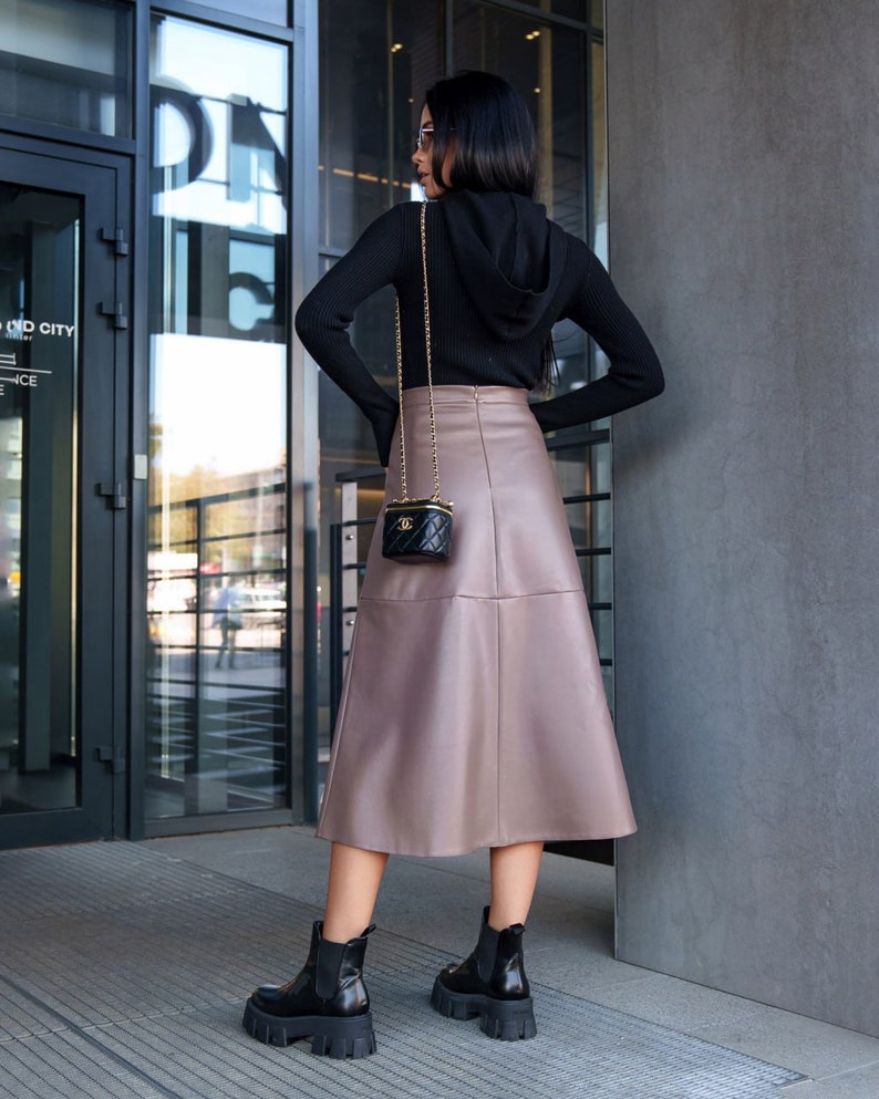 Pu&ograve; includere: Una donna indossa un maglione nero con cappuccio e una gonna di pelle marrone. Porta una piccola borsa nera con una tracolla a catena dorata. Indossa stivaletti neri.