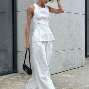 May include: A woman wearing a white sleeveless top with a criss-cross design and matching white wide-leg pants. She is carrying a black handbag and wearing black sandals.