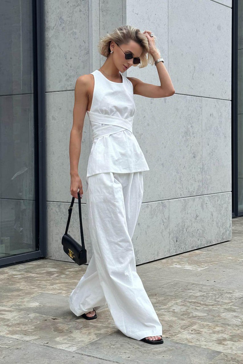 May include: A woman wearing a white sleeveless top with a criss-cross design and matching white wide-leg pants. She is carrying a black handbag and wearing black sandals.