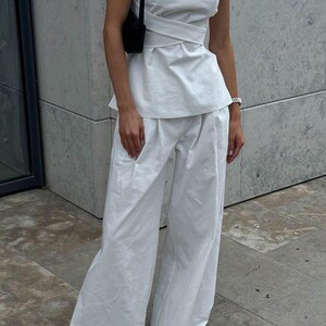 May include: A white sleeveless top with a wrap-style design and a white pair of wide-leg pants. The top has a round neckline and a fitted bodice. The pants have a high waist and a relaxed fit.