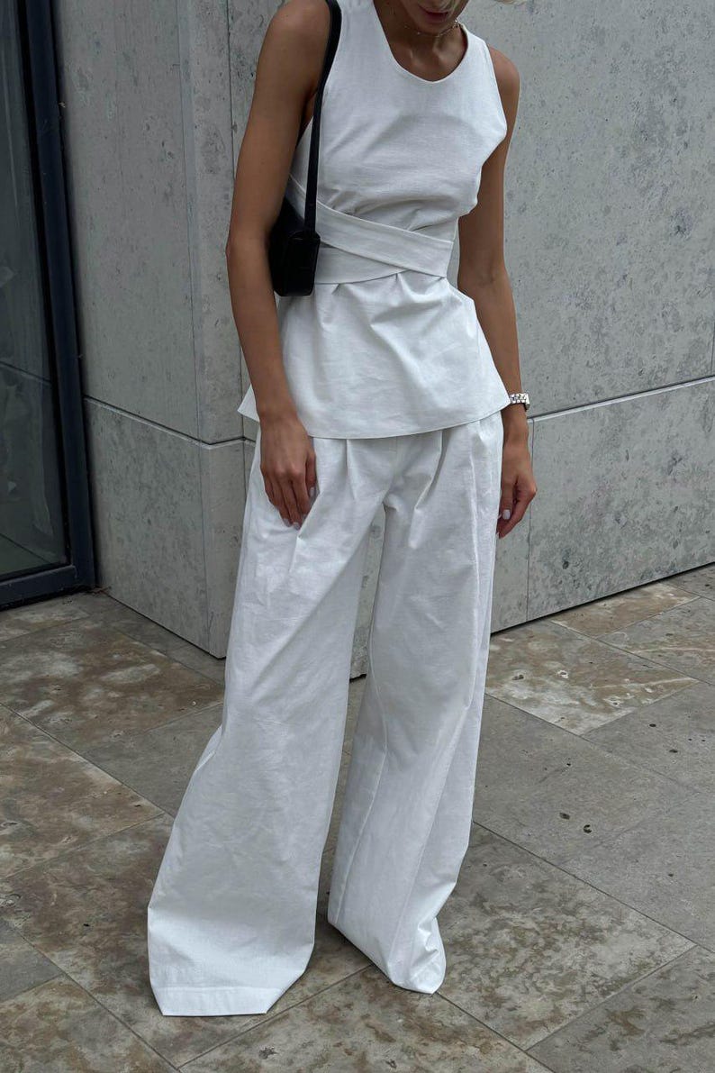 May include: A white sleeveless top with a wrap-style design and a white pair of wide-leg pants. The top has a round neckline and a fitted bodice. The pants have a high waist and a relaxed fit.