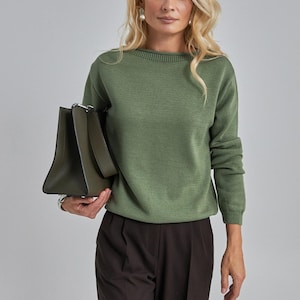 Op de afbeelding: Een vrouw draagt een groene trui en een donkerbruine broek en houdt een donkergroene handtas vast. De trui heeft een ronde hals en lange mouwen. De broek is op maat gemaakt. De vrouw heeft blond haar en draagt oorbellen.