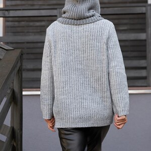 May include: A gray hooded sweater with a ribbed knit texture. The sweater has a high collar and a hood that covers the head. The person is wearing black leather pants. The background is a wooden wall and a gray wall.
