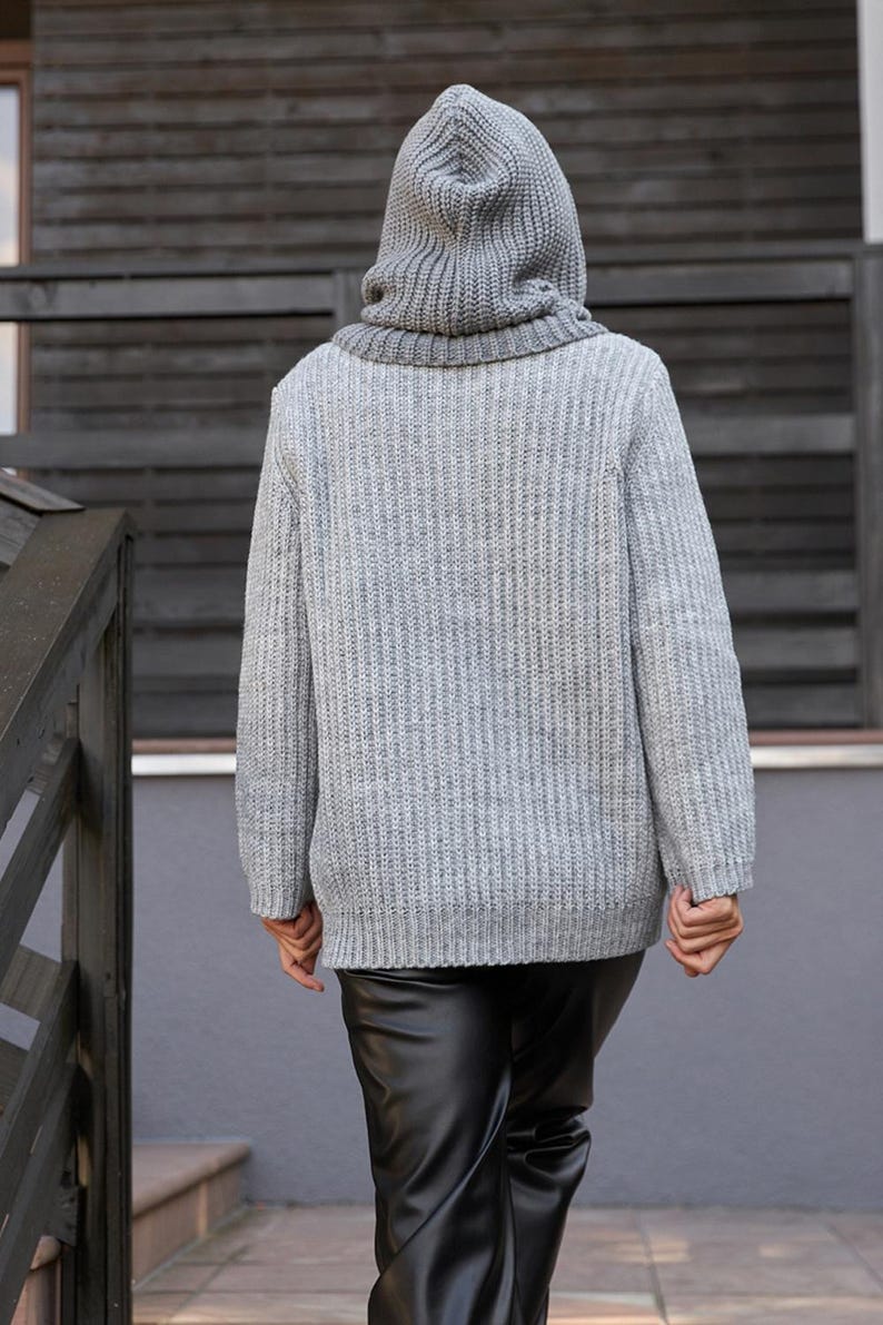 May include: A gray hooded sweater with a ribbed knit texture. The sweater has a high collar and a hood that covers the head. The person is wearing black leather pants. The background is a wooden wall and a gray wall.