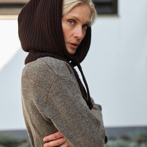 May include: A woman wearing a brown knit hood and a long-sleeved brown coat. The hood is a darker shade of brown and is tied with drawstrings. The coat is cinched at the waist with a brown belt.