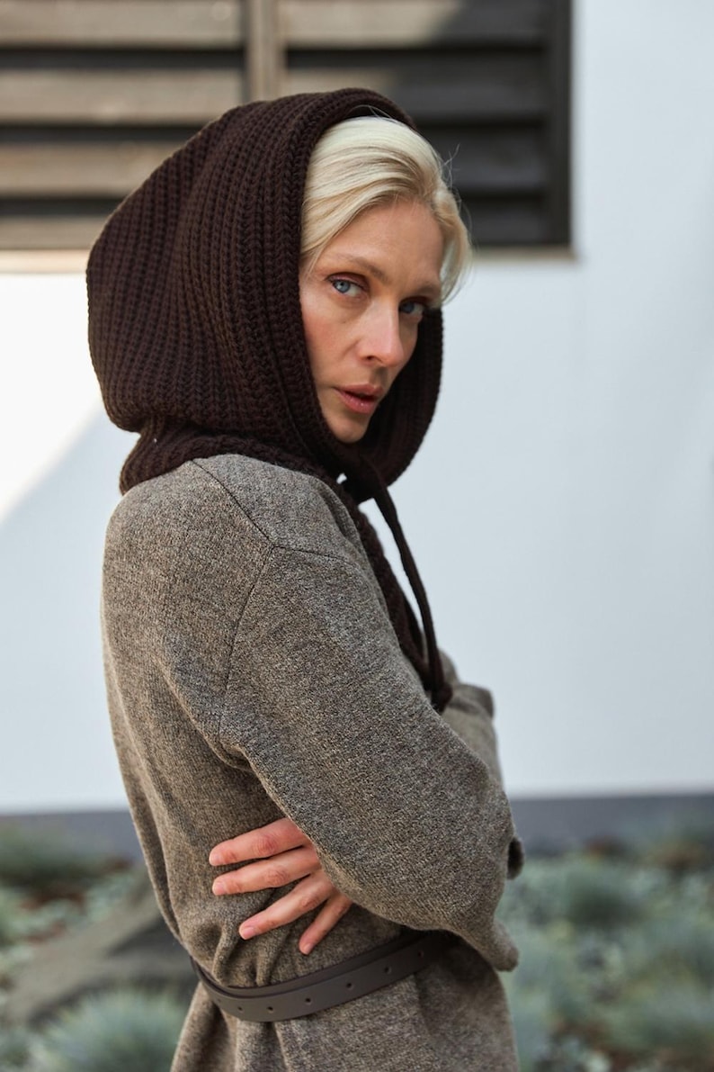 May include: A woman wearing a brown knit hood and a long-sleeved brown coat. The hood is a darker shade of brown and is tied with drawstrings. The coat is cinched at the waist with a brown belt.