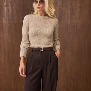 May include: A woman wearing a beige ribbed sweater, dark brown pleated trousers, and a brown belt with a silver buckle. She is also wearing sunglasses. The background is a textured brown wall.