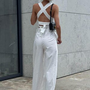 May include: A white crop top with a criss-cross back and a bow tie detail. The top is paired with white wide-leg pants. The model is wearing a small black purse.