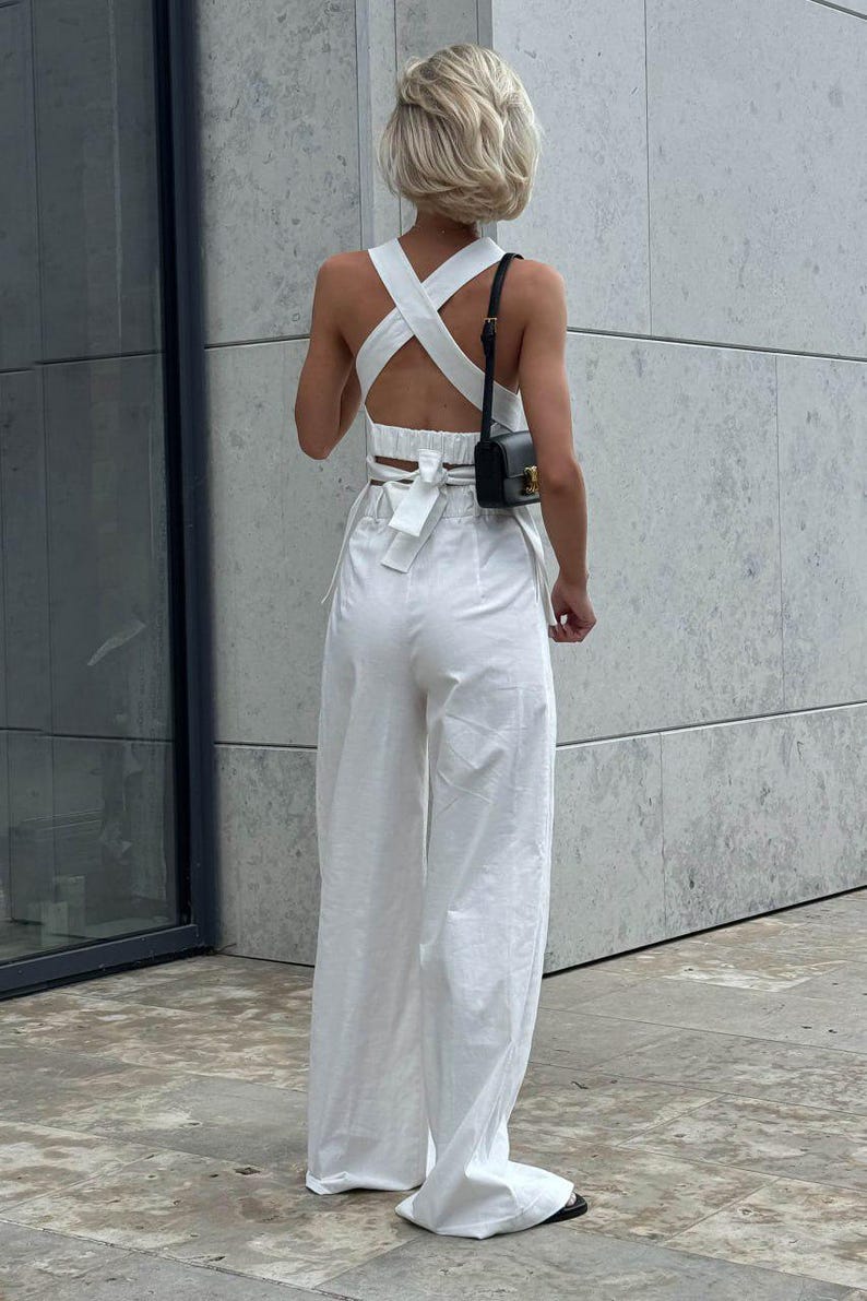 May include: A white crop top with a criss-cross back and a bow tie detail. The top is paired with white wide-leg pants. The model is wearing a small black purse.