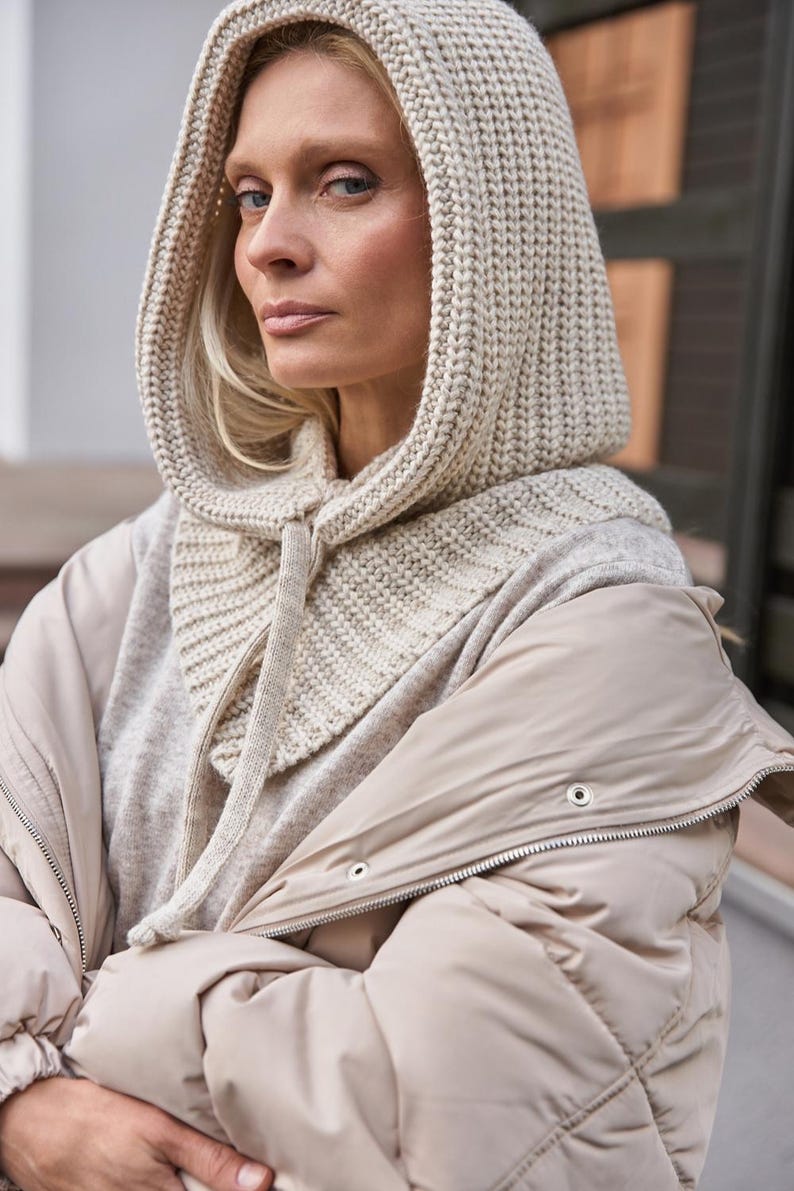 May include: A beige knit hood and scarf combination with a drawstring closure. The hood is worn over a light gray sweater and a beige puffer jacket. The outfit is neutral-toned and suitable for cold weather.