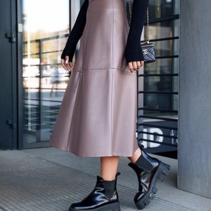Pu&ograve; includere: Una donna indossa una gonna midi in similpelle marrone con un maglione nero e stivaletti neri.