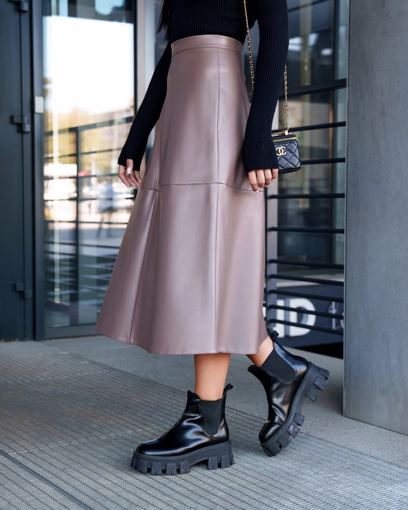 Pu&ograve; includere: Una donna indossa una gonna midi in similpelle marrone con un maglione nero e stivaletti neri.
