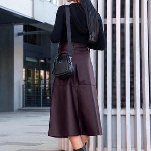 Pu&ograve; includere: Una donna indossa un maglione nero, una gonna in similpelle bordeaux e stivaletti neri. Porta una piccola borsa a mano nera con una tracolla.