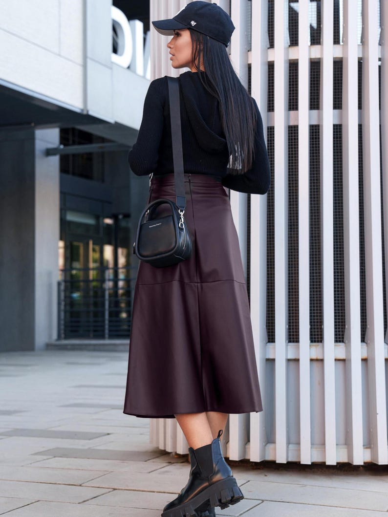 Pu&ograve; includere: Una donna indossa un maglione nero, una gonna in similpelle bordeaux e stivaletti neri. Porta una piccola borsa a mano nera con una tracolla.